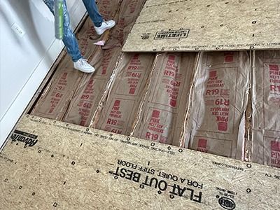 A worker installs subflooring over floor joists insulated with R-19 fiberglass batts.
