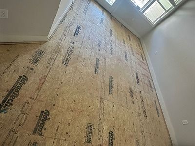 A room under construction with bare Advantech subflooring and white walls, looking toward a windowed exterior door.