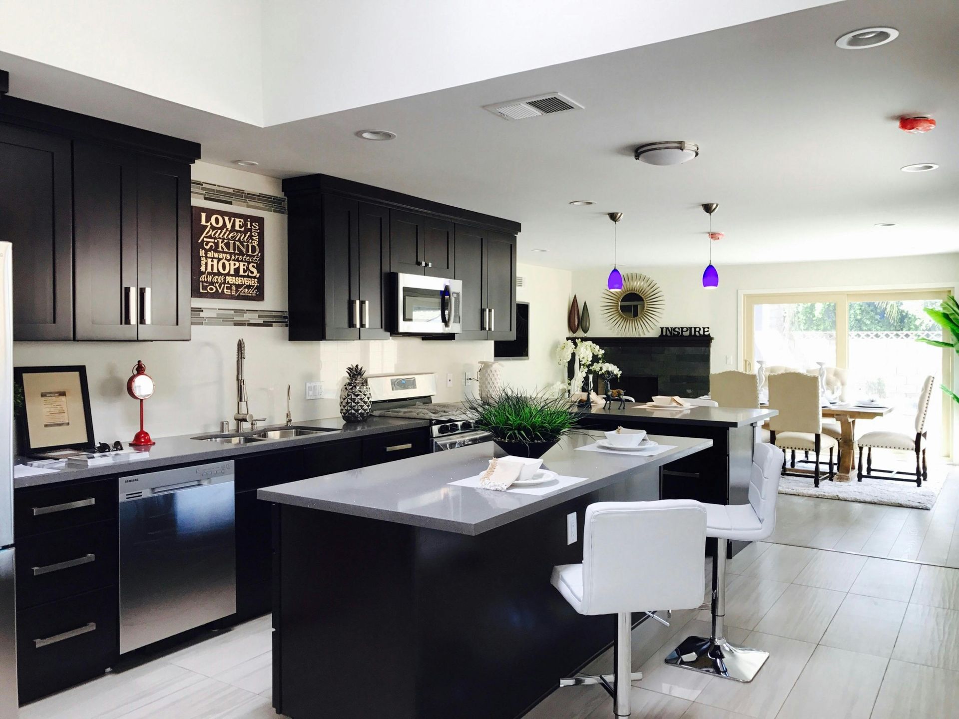 Modern kitchen with dark cabinets, a large island with white stools, and light-colored flooring, leading to a dining area.
