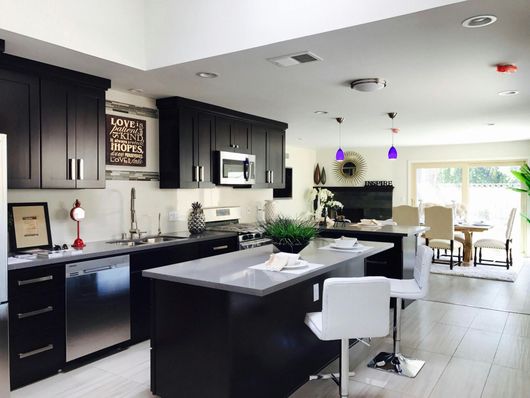 Modern kitchen with dark cabinets, a large island with white stools, and light-colored flooring, leading to a dining area.