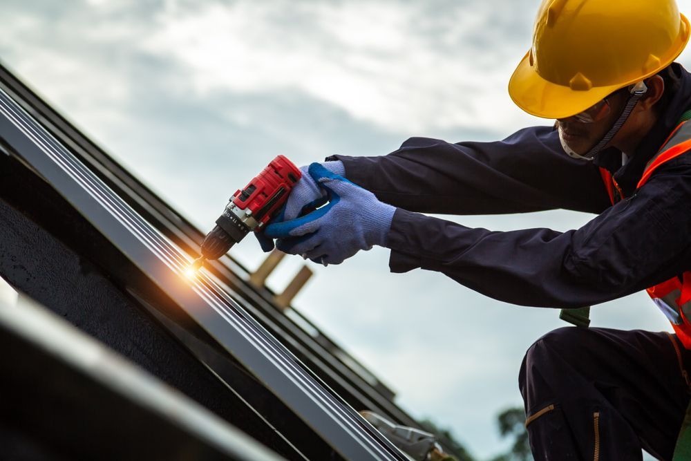 A construction worker wearing a yellow hard hat and orange safety vest uses a power drill on a metal roof.