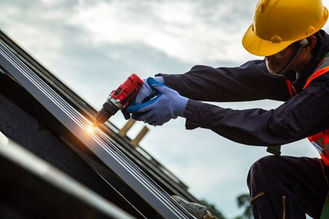 A construction worker wearing a yellow hard hat and orange safety vest uses a power drill on a metal roof.
