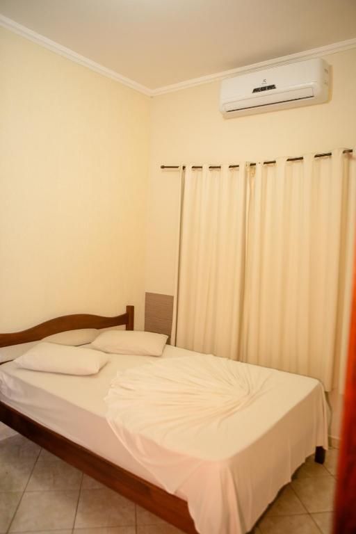 Um quarto com uma cama de madeira, roupa de cama branca, cortinas bege cobrindo uma janela e um ar-condicionado de parede.