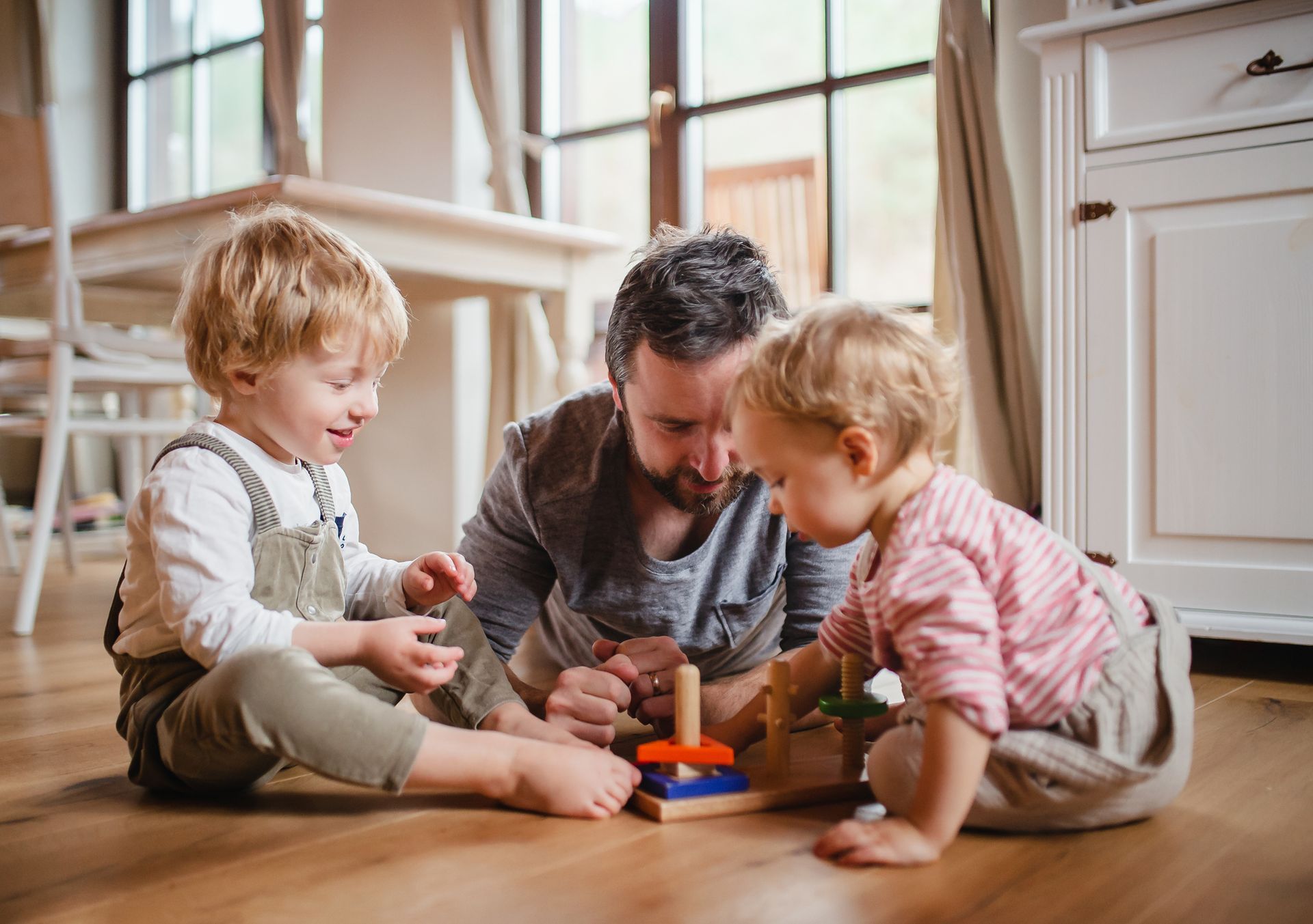 A parent and two children sit on a wooden floor, focused on playing with a colorful wooden stacking toy.