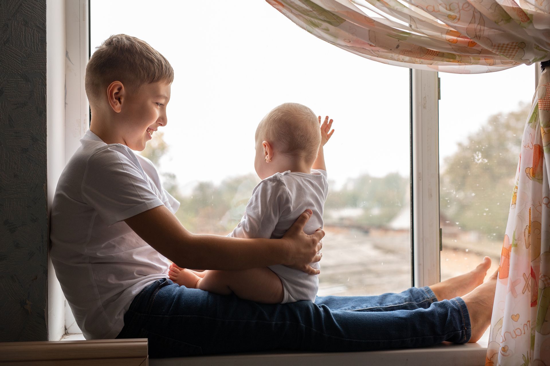 A child sitting on a windowsill, gently holding a baby who is reaching toward the window.