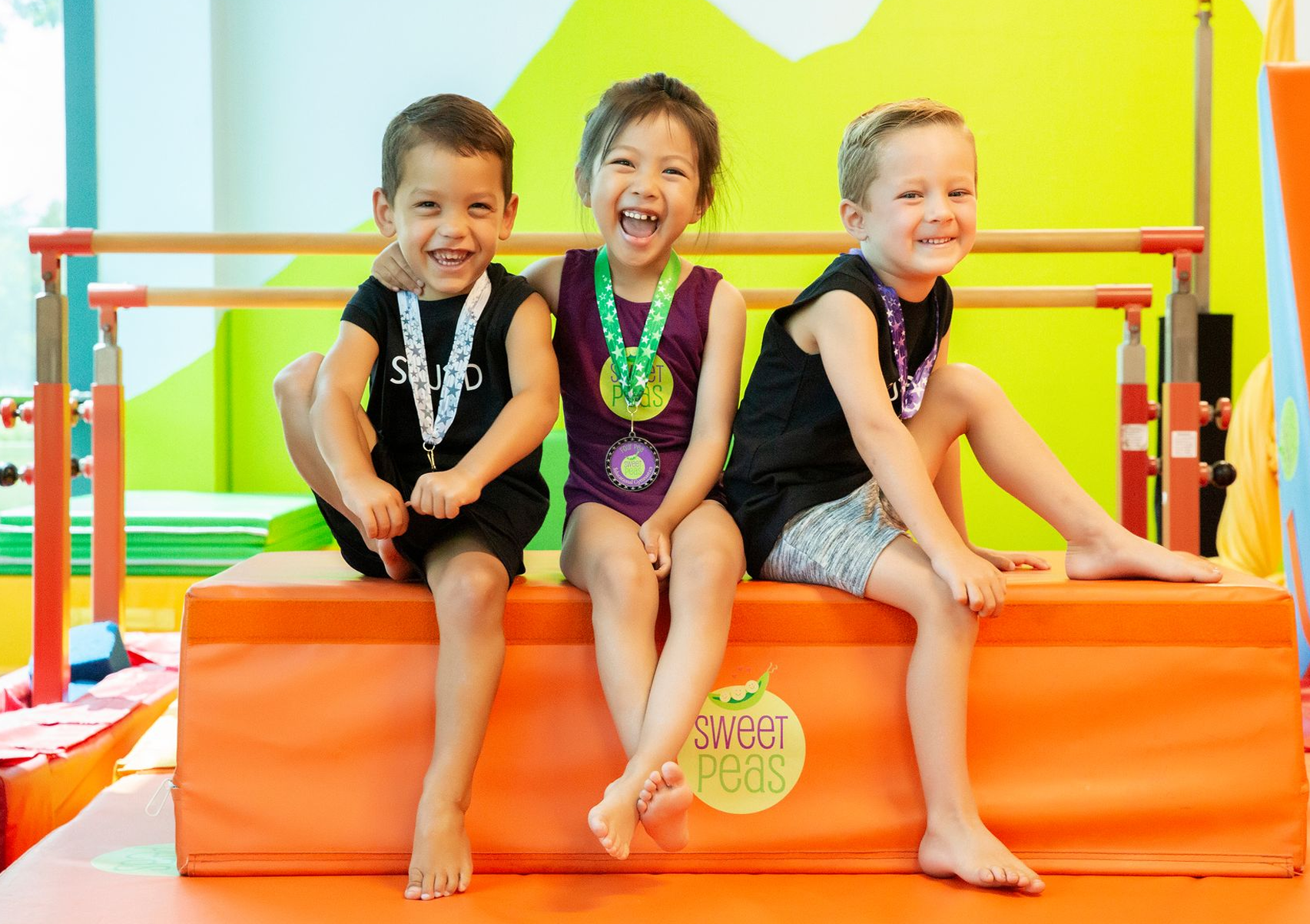Three young children are sitting on a mat in a gym.