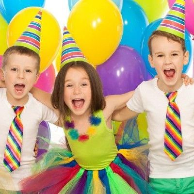 Three children wearing party hats and ties are standing in front of balloons.