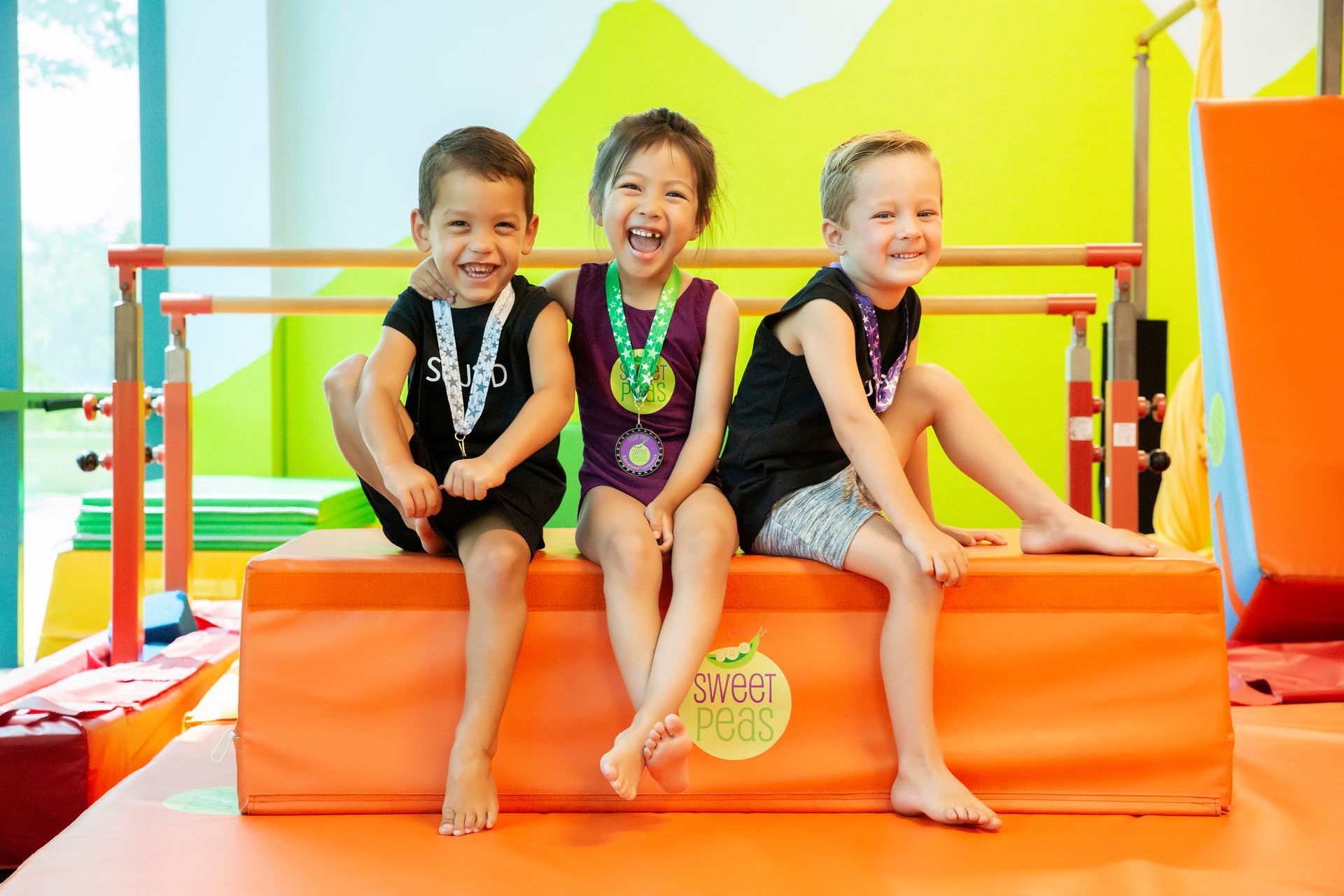 Three young children are sitting on a mat in a gym.