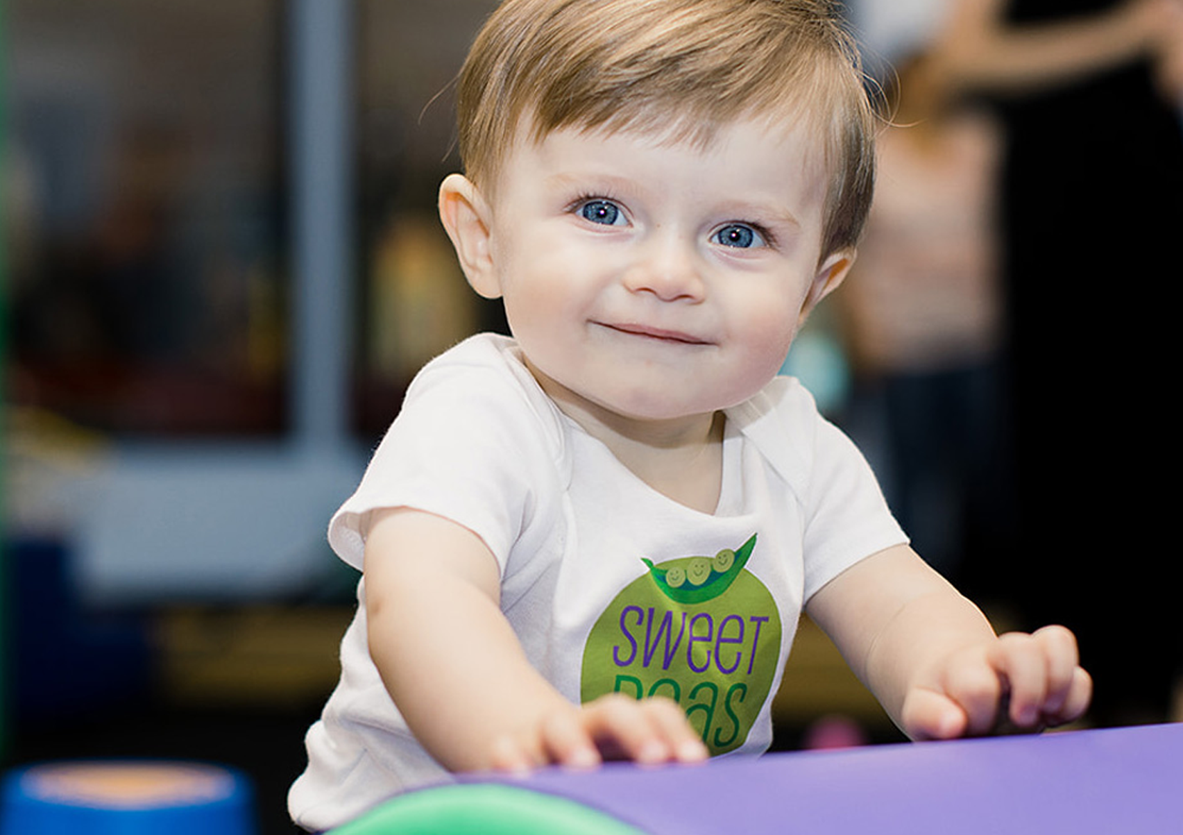 A baby is sitting on a purple block and looking at the camera.