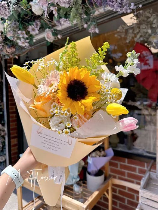 户外手持花束，由向日葵、白色和黄色花朵组成，用米色纸包裹，花束内容为向日葵、白色和黄色花朵。
