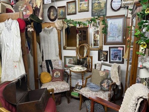 Vintage shop interior with white garments, framed art, mirrors, and assorted antique furniture