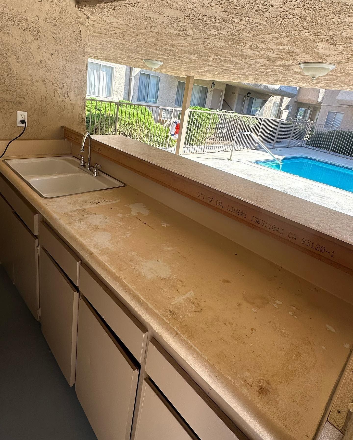 Stone countertop and sink beside a window overlooking a blue swimming pool and patio.