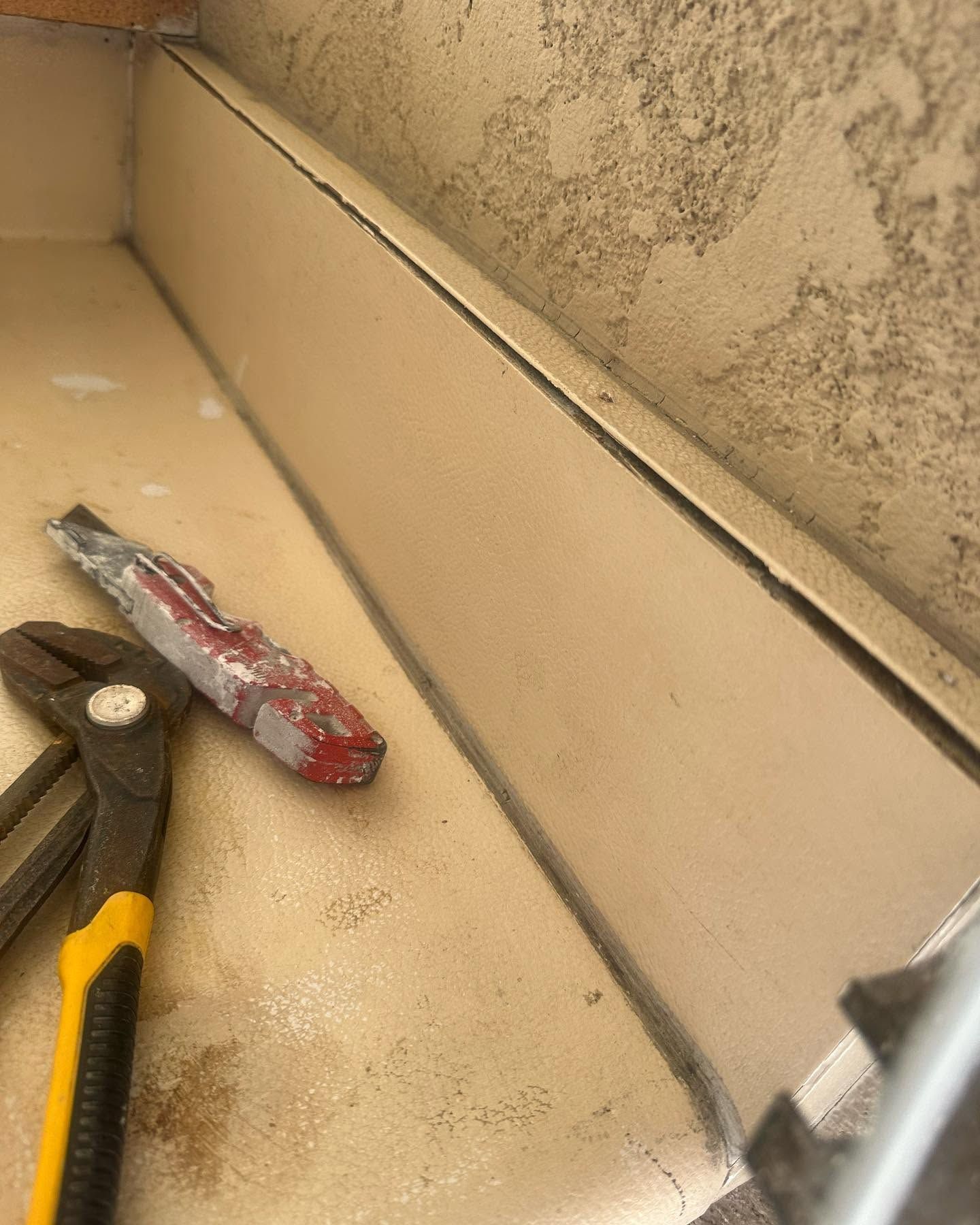 Pliers and a red-handled tool on a dusty beige ledge beside a textured wall.