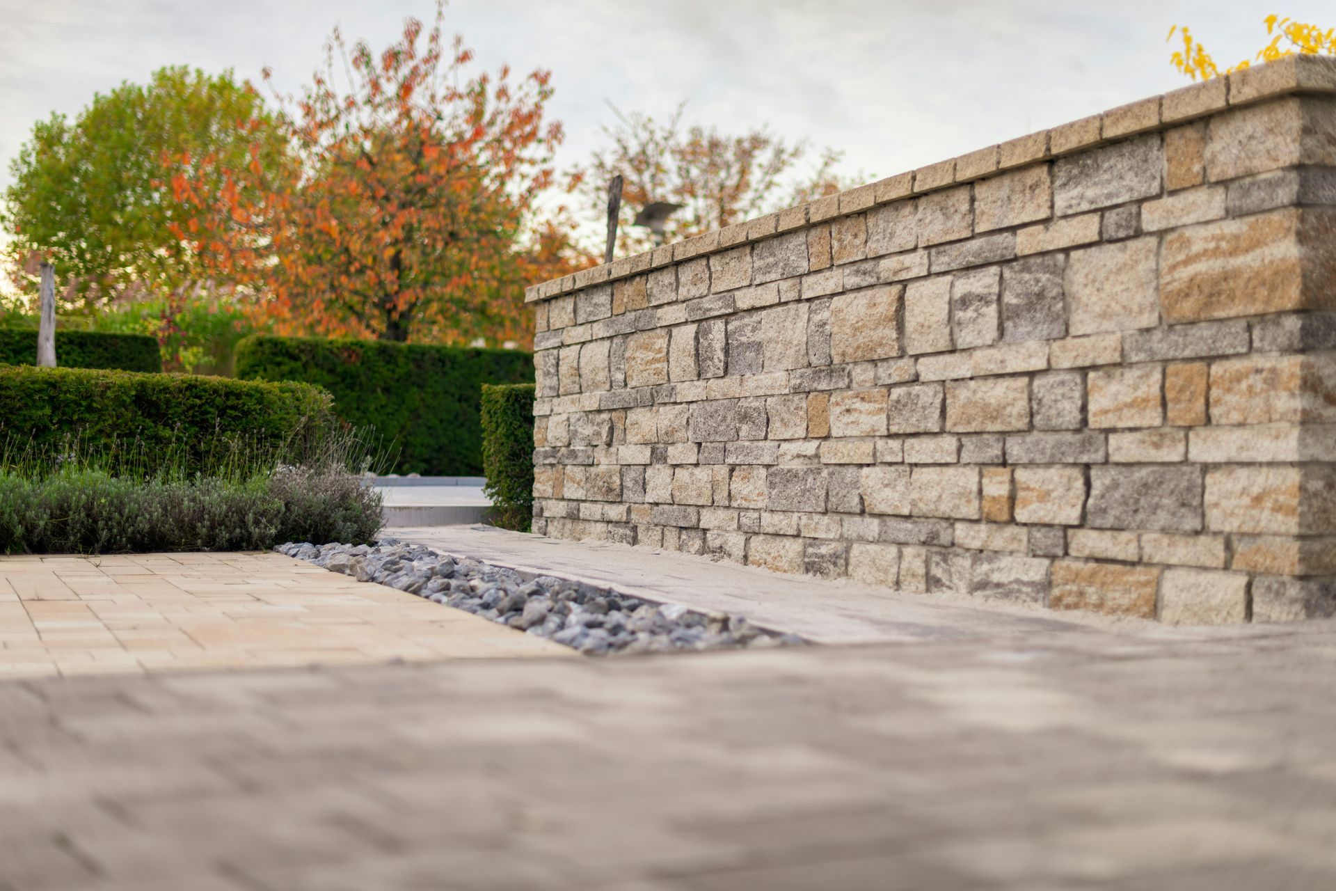 Stone retaining wall beside a landscaped walkway with hedges and autumn trees