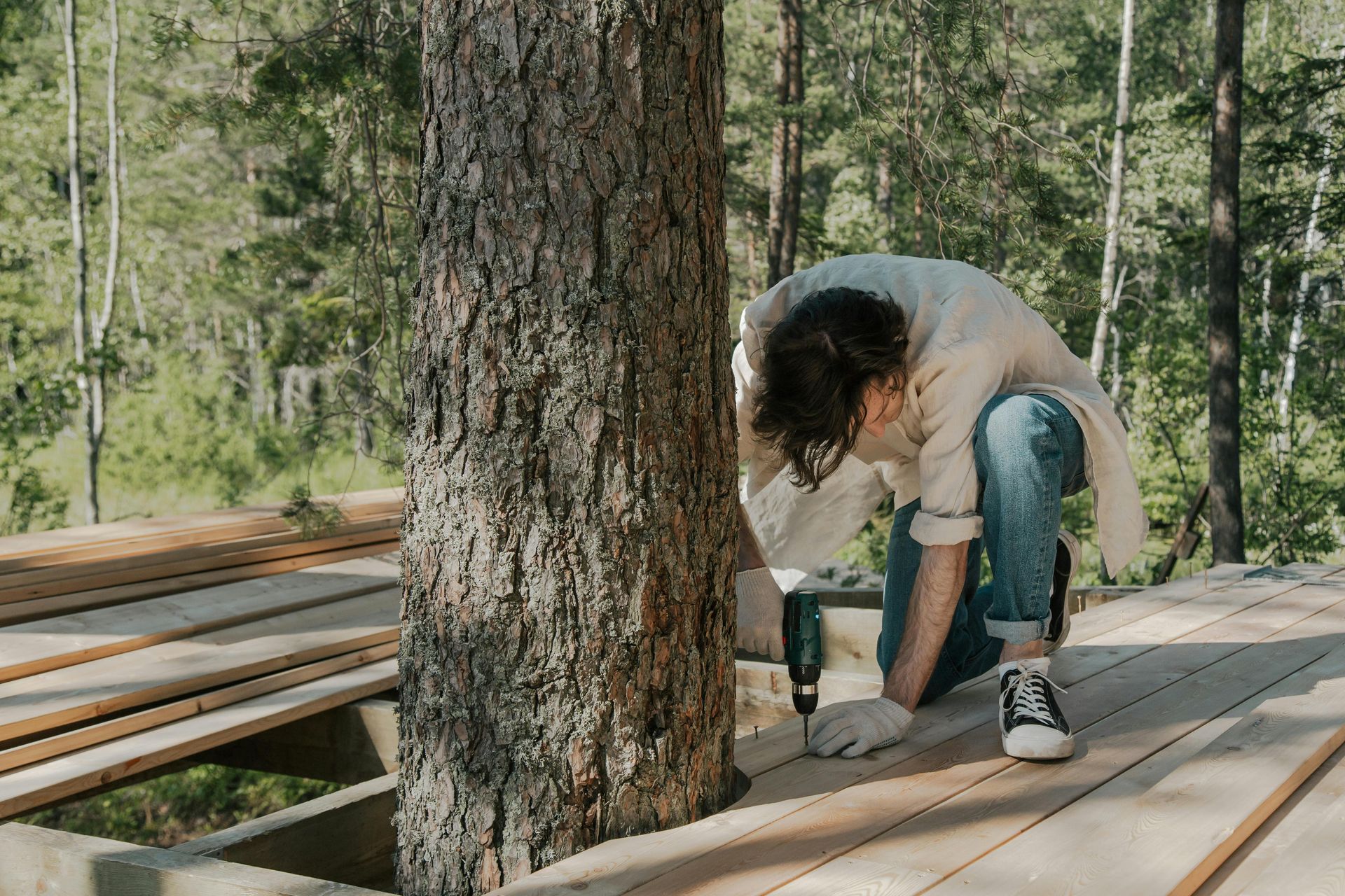 Person using a power drill to build a wooden deck around a tree outdoors.
