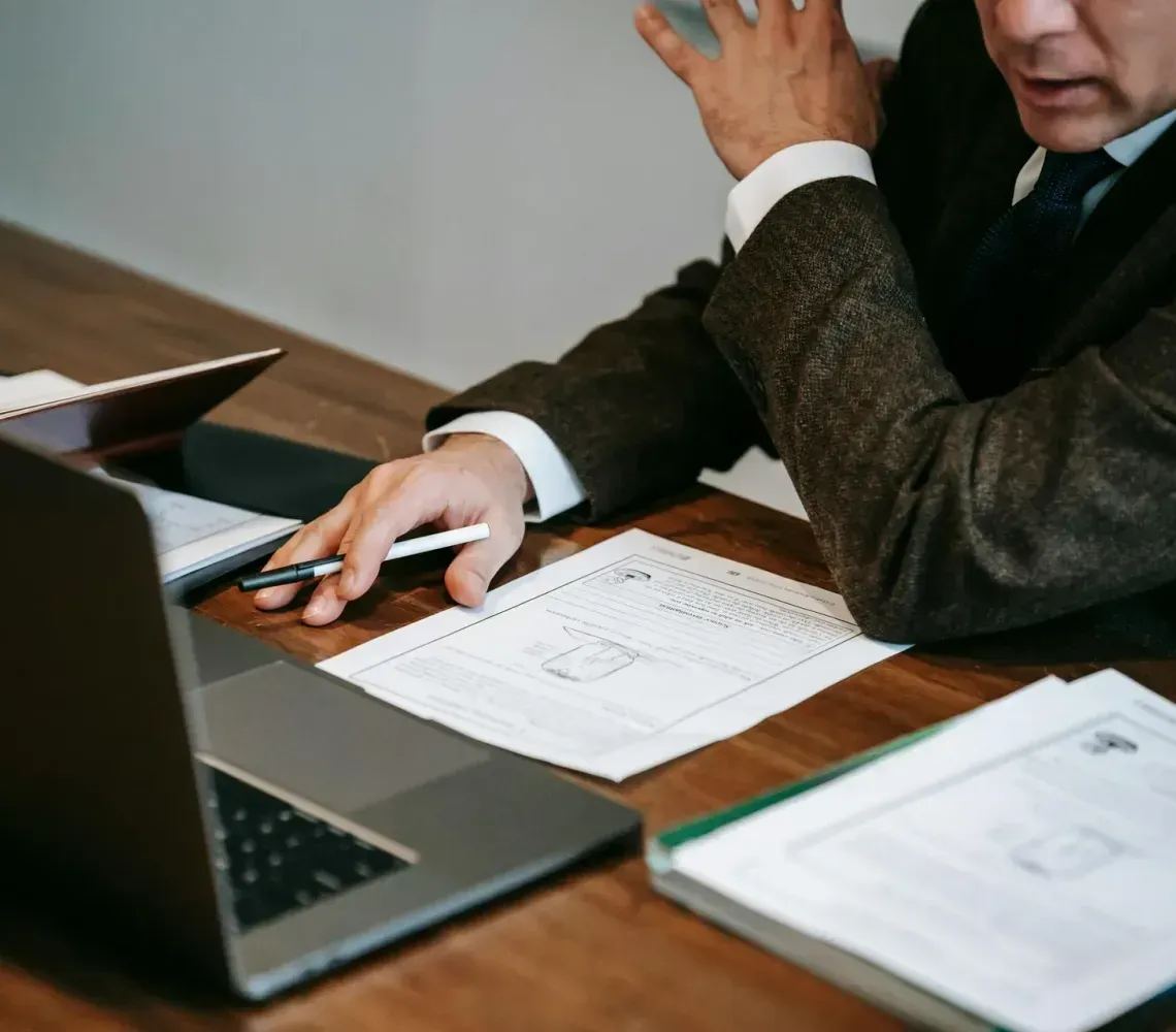 Person in a dark suit reviewing documents at a desk with a laptop and notebook