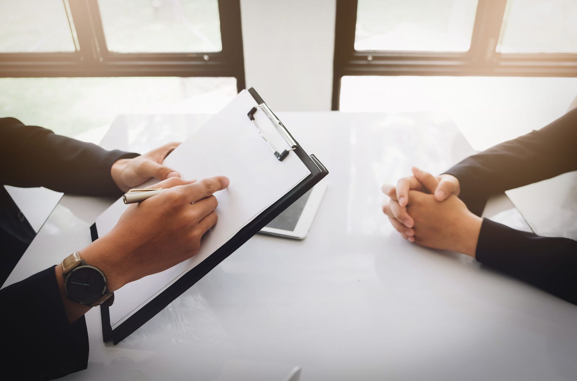 Two people in a business meeting, one taking notes on a clipboard across a white table by a bright window.