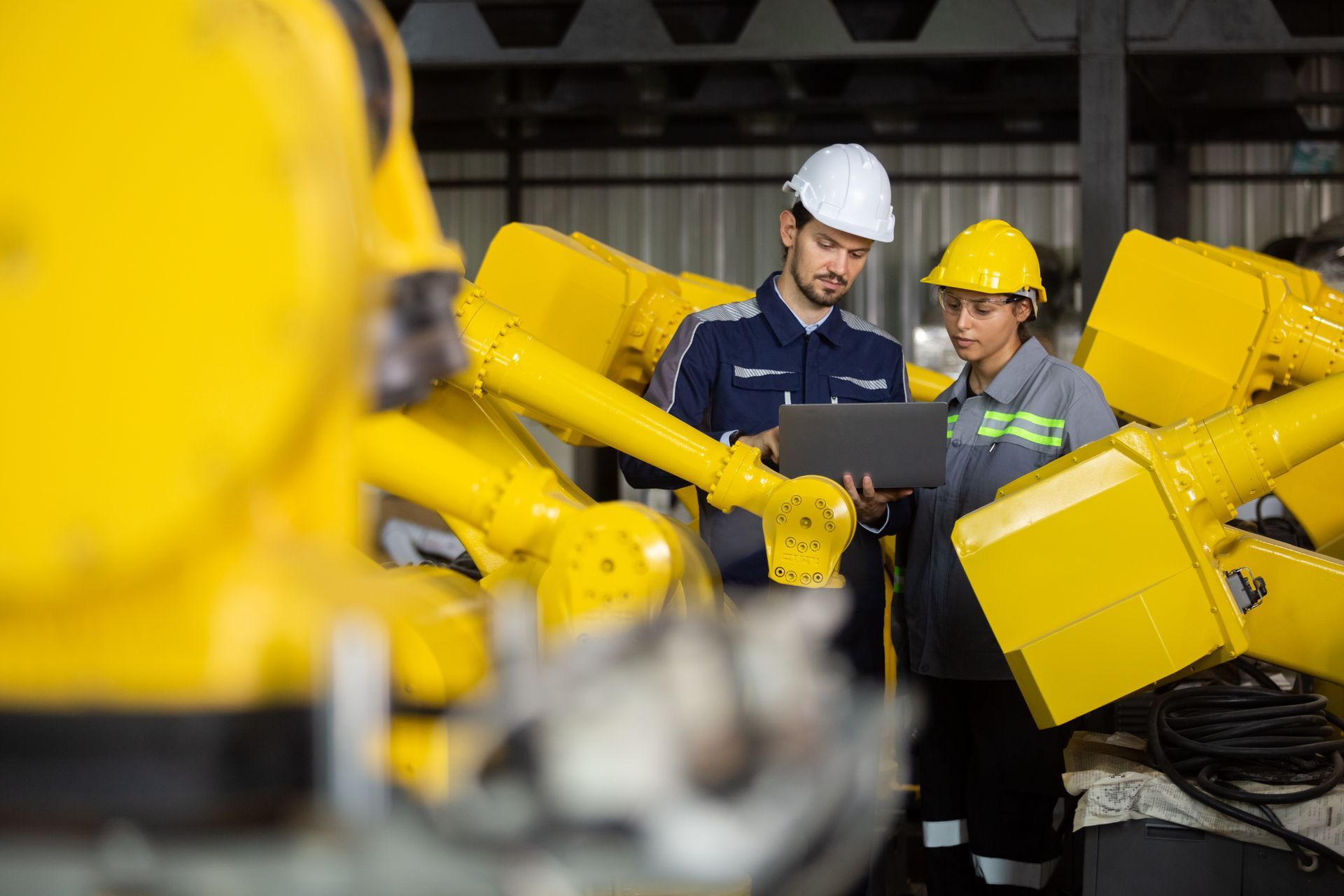 Two engineers in hard hats reviewing a tablet beside bright yellow industrial machinery