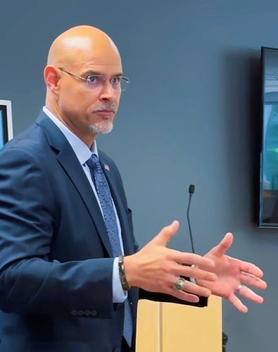 Man in a navy suit gesturing while speaking in a conference room, beside a wall-mounted screen.