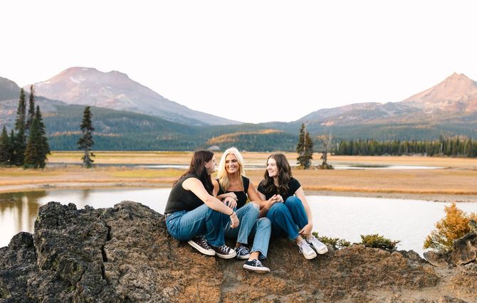 Three women are sitting on top of a rock near a lake.