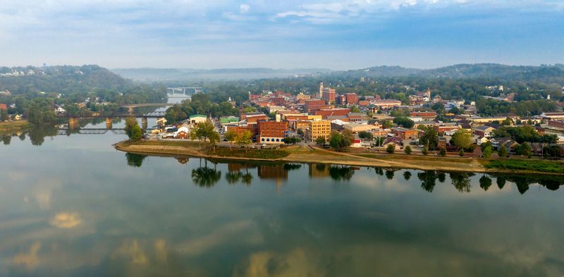 Aerial view of a lakeside town with colorful buildings, trees, and hills reflected in calm water.