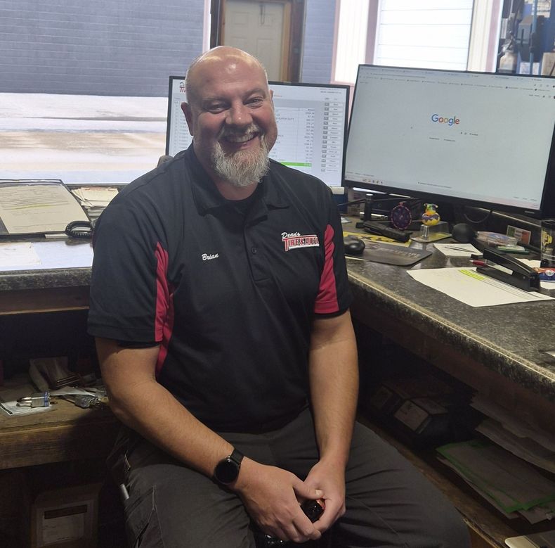 Smiling man seated at a desk with dual computer monitors in an office. | Dean's Tire & Auto