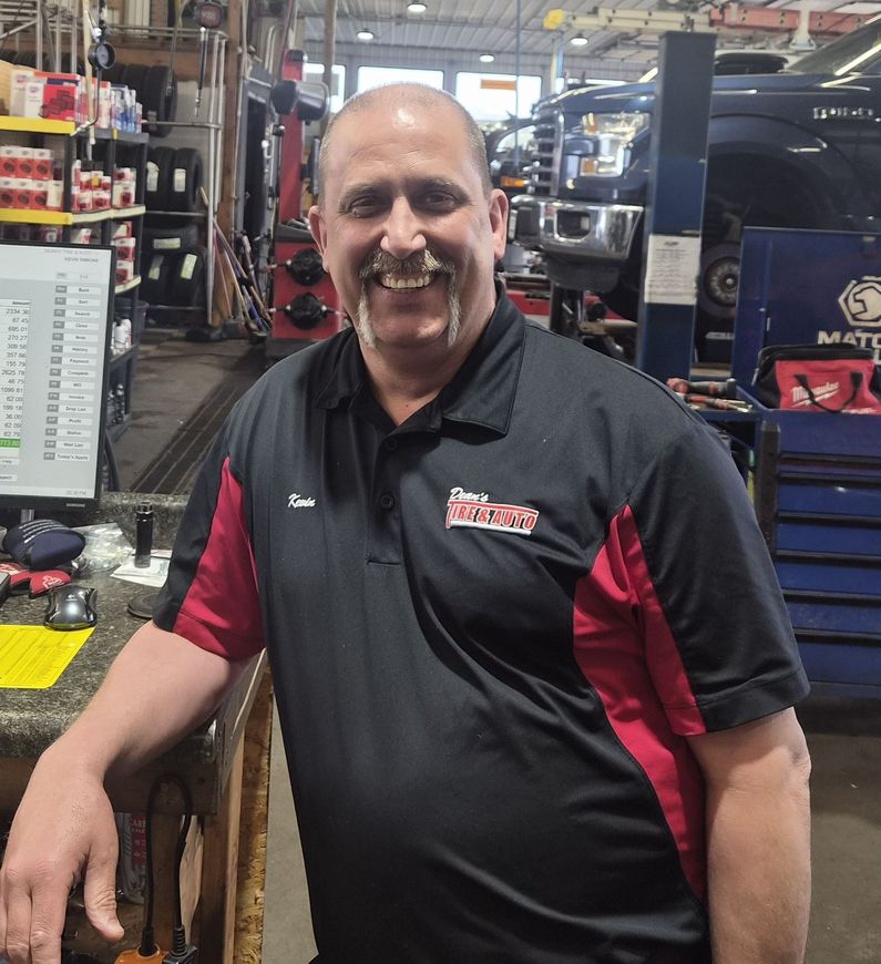 Mechanic smiling in a auto repair shop, wearing a black-and-red work shirt. | Dean's Tire & Auto