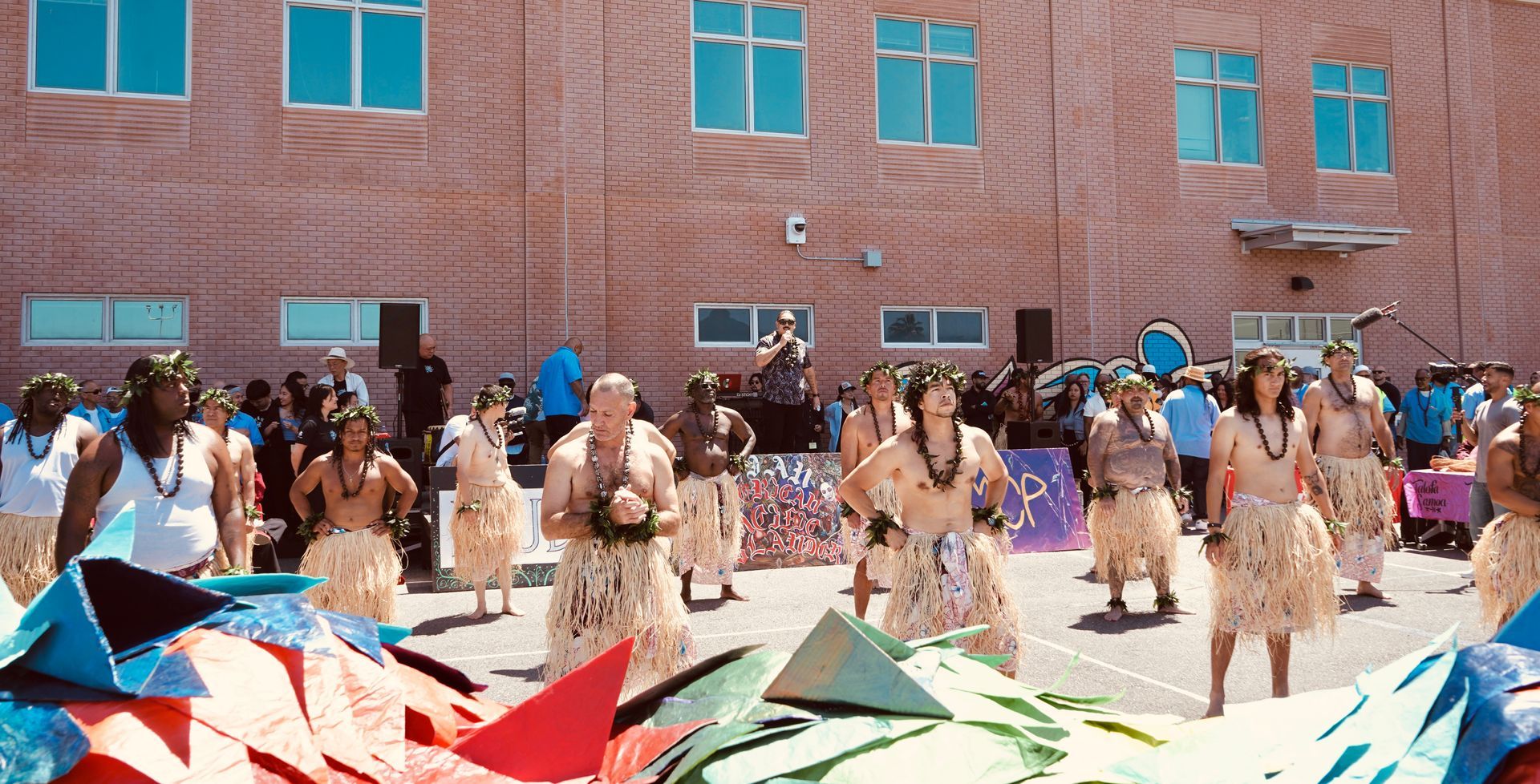 People gathered outdoors in front of a brick building, with colorful kites or fabric spread in the foreground.