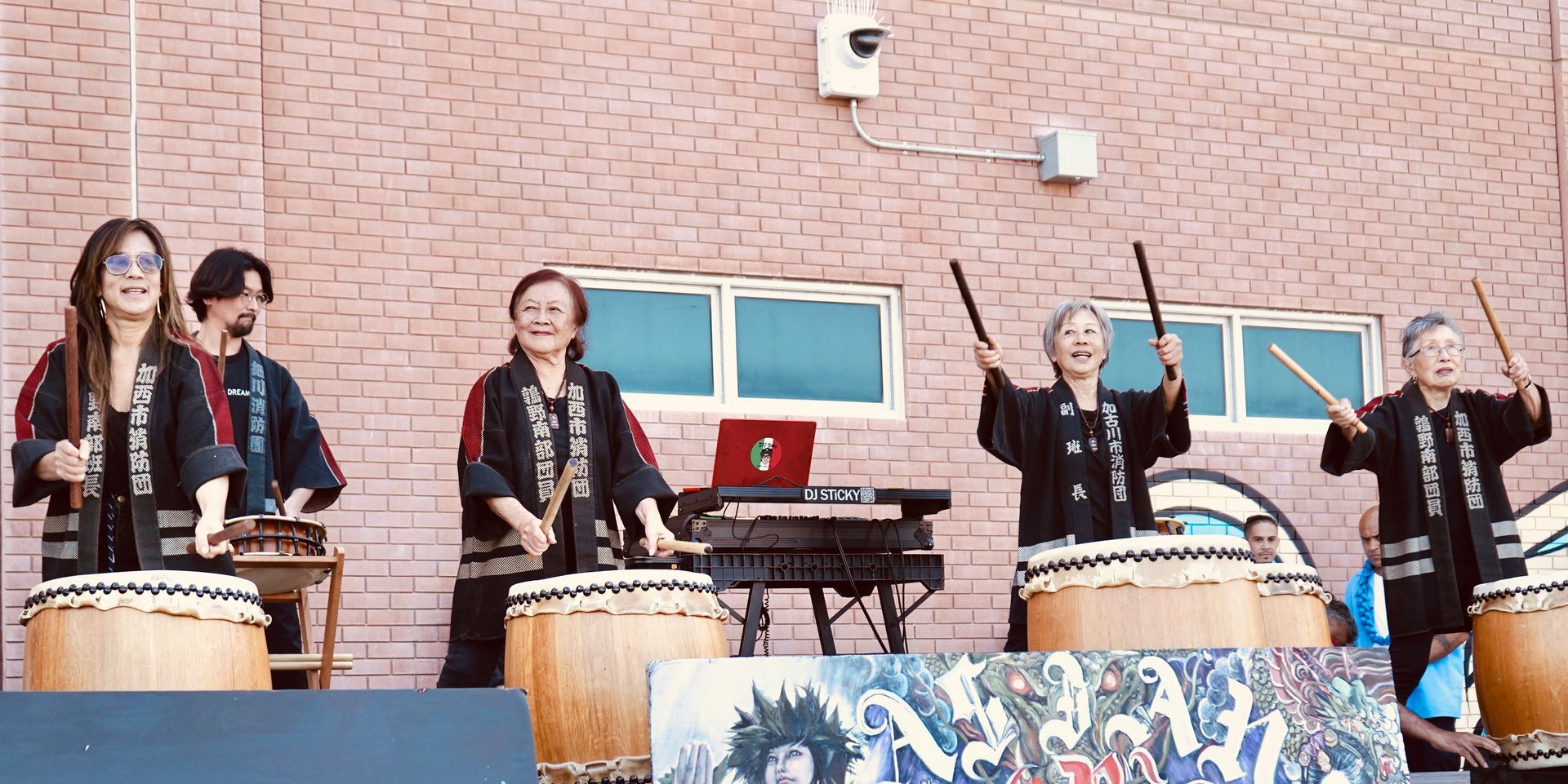 Drummers performing outdoors against a brick wall, striking large drums and a keyboard setup behind them