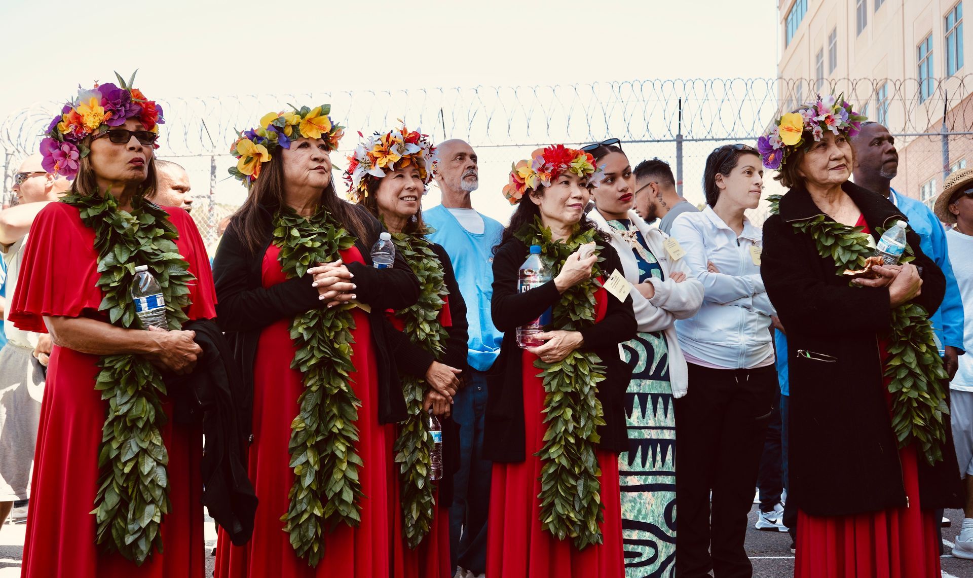 Women in red dresses and flower crowns stand outdoors holding leafy leis in a group performance