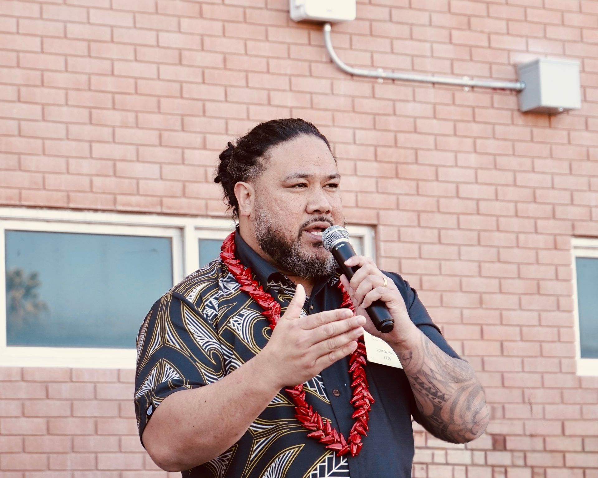 Man speaking into a microphone outdoors, wearing a patterned shirt and red lei against a brick wall.