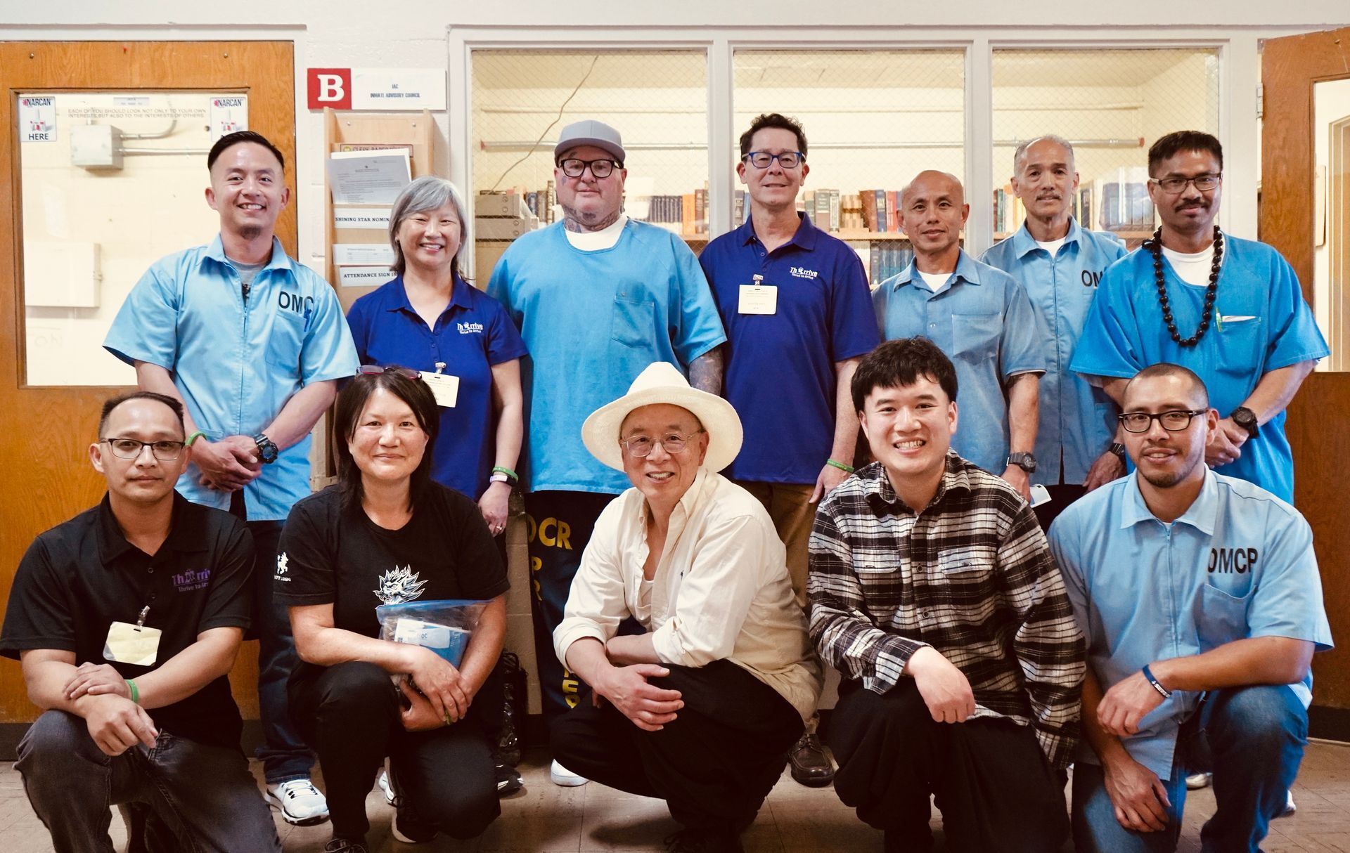 Group of smiling people in blue shirts posing indoors in front of windows and a bulletin board.