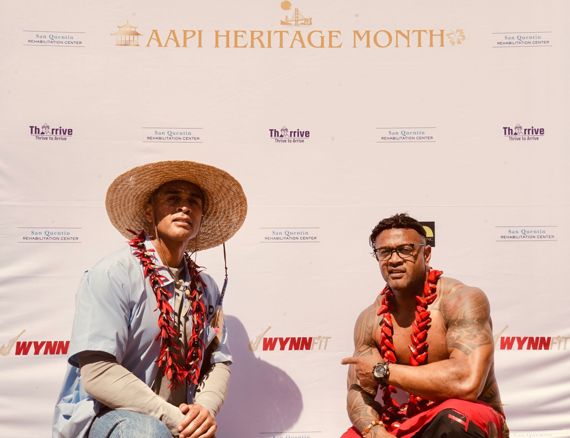 Two people posing at an AAPI Heritage Month step-and-repeat backdrop, one in a straw hat and one shirtless with a lei.