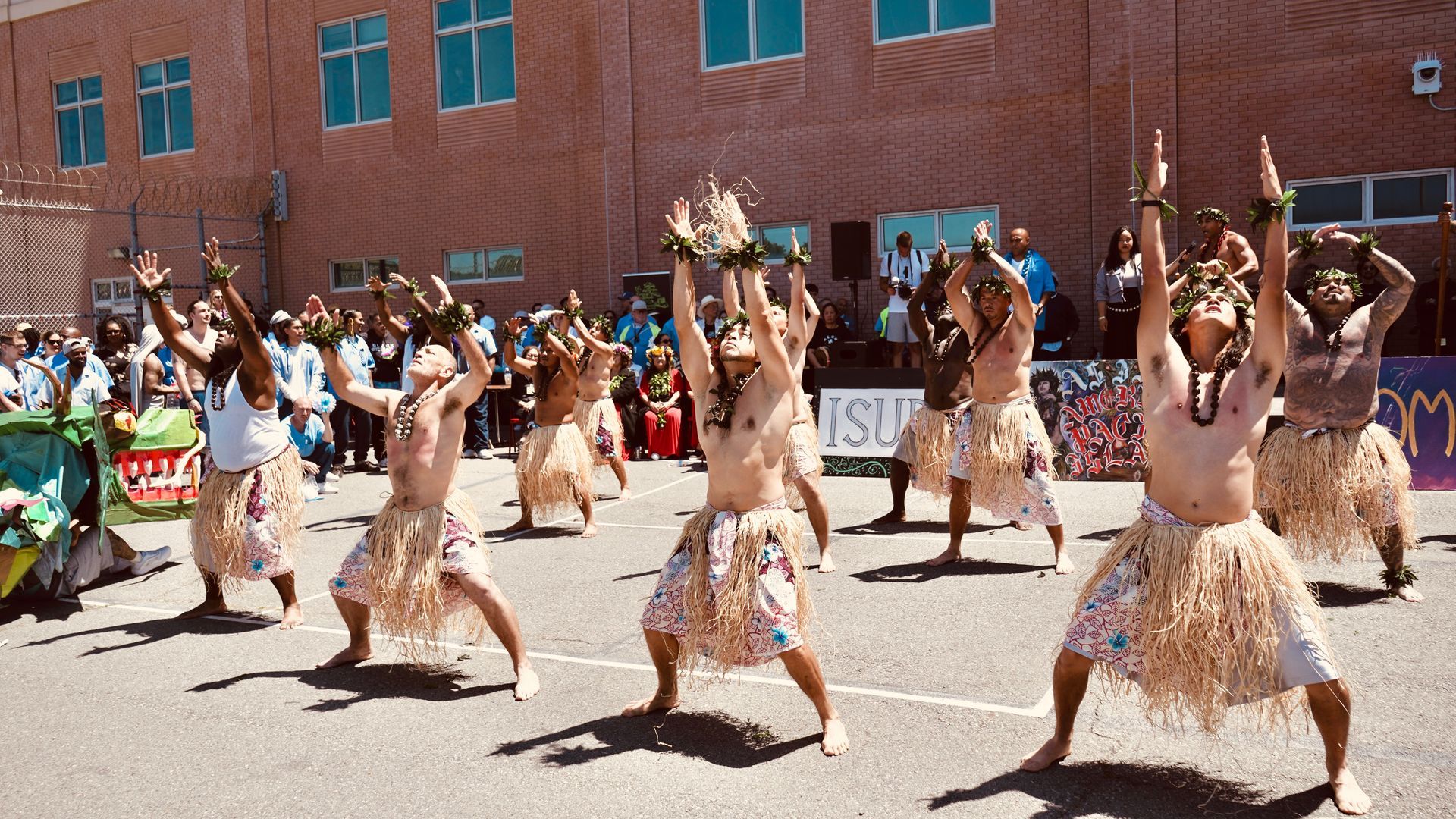 Dancers in patterned skirts perform outdoors in front of a brick building, arms raised in synchronized movement