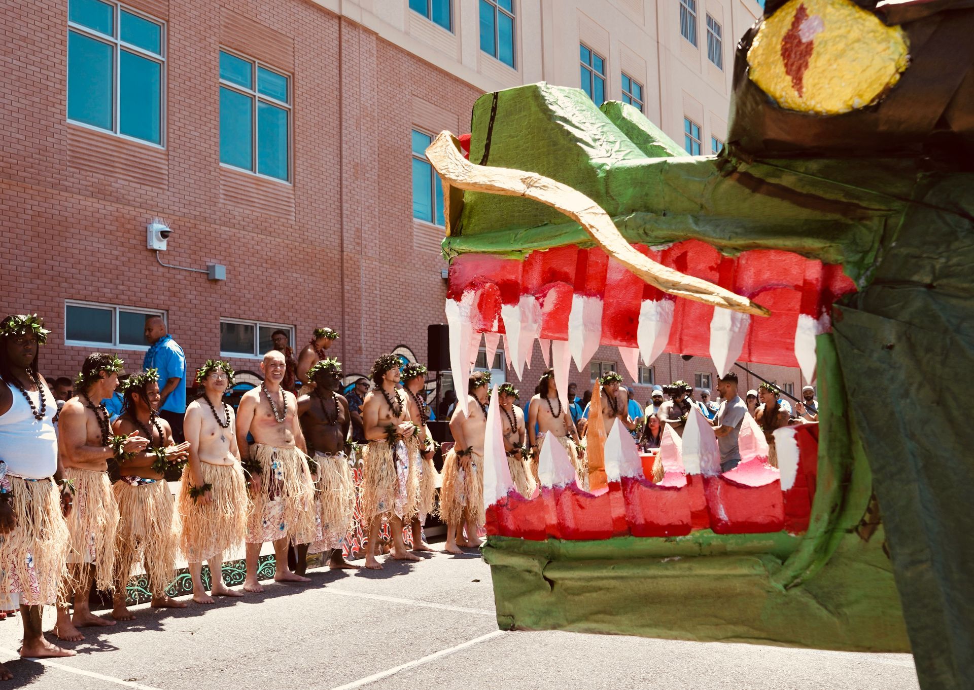 People in traditional attire line a street beside a large red, green, and white parade float.