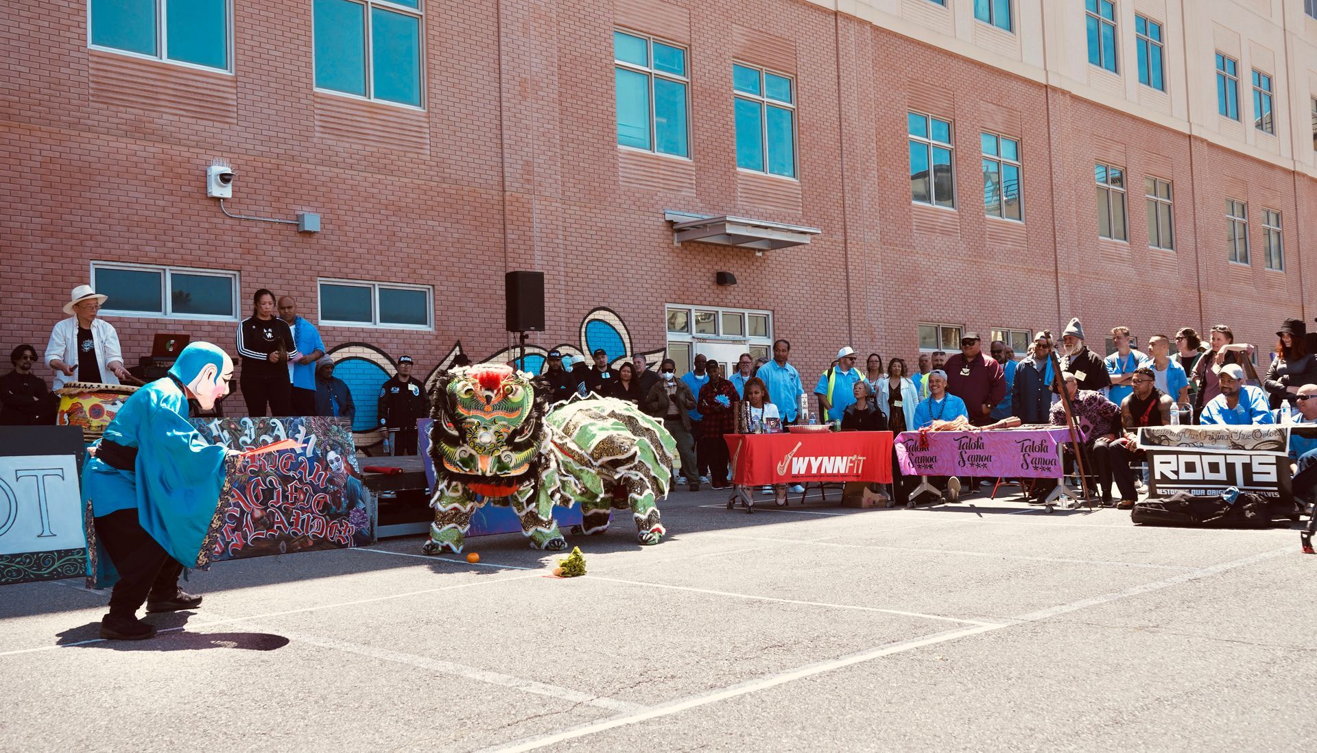 Outdoor crowd watches two lion dancers performing on a street in front of a brick building.