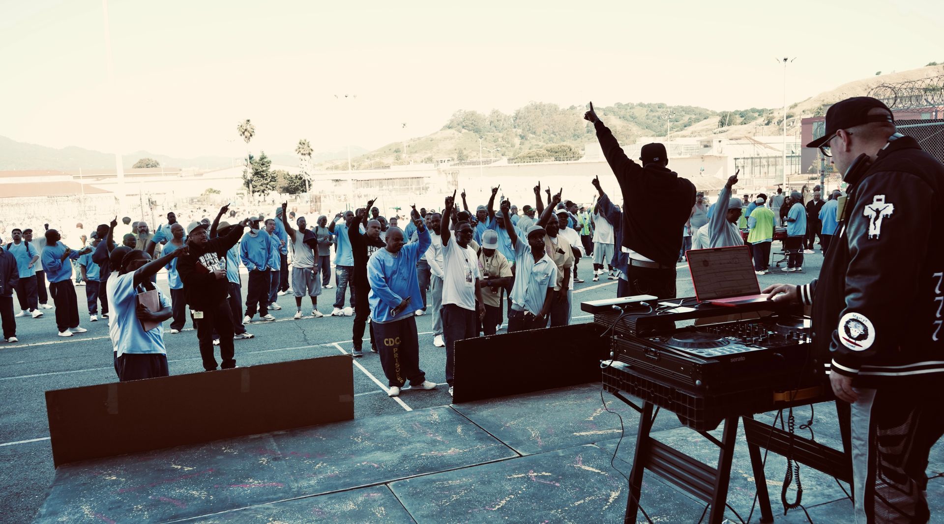 DJ performing for a large outdoor crowd with raised hands at a sunny beach event