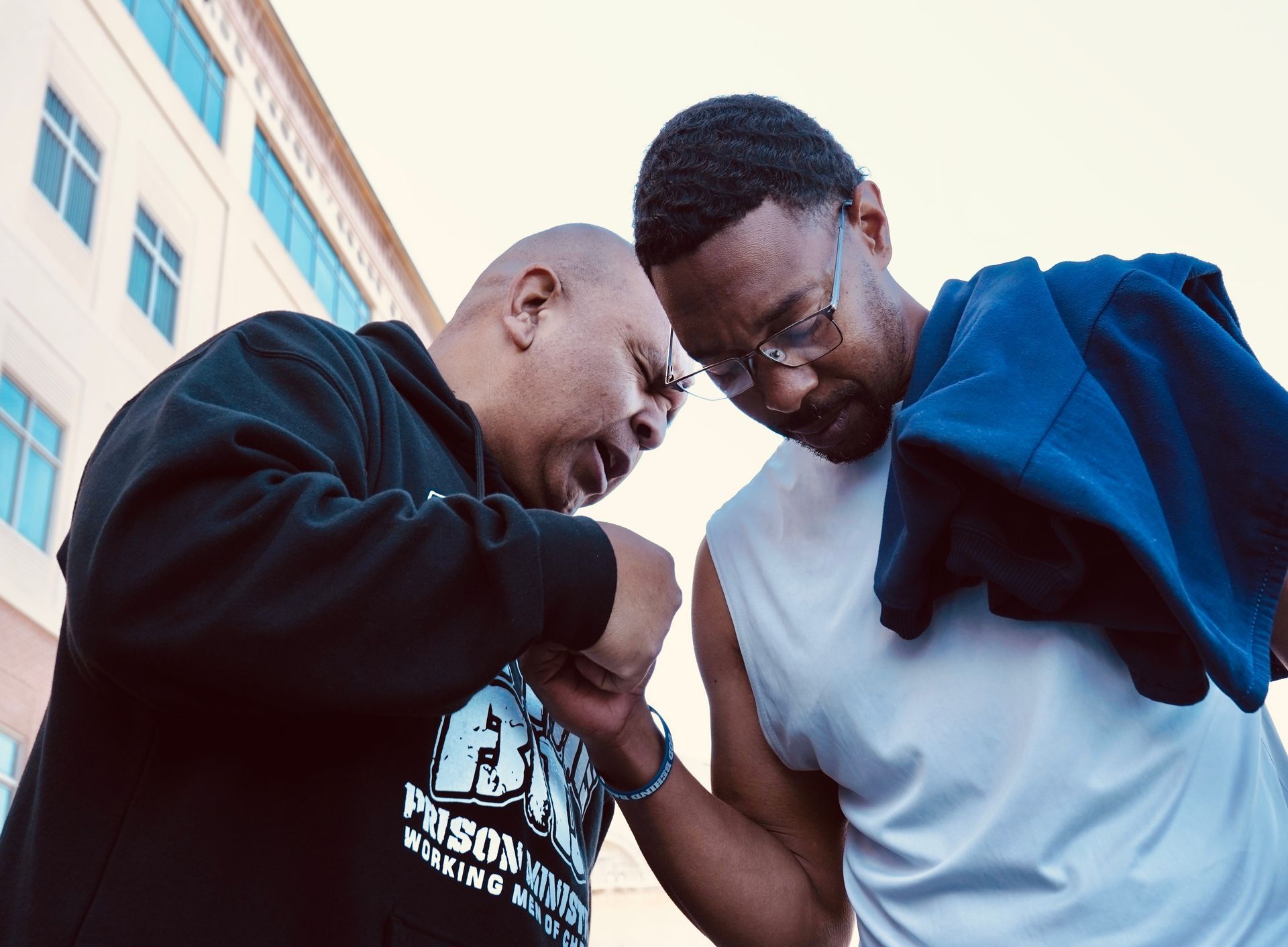 Two people leaning in outdoors, smiling and touching fists in front of a building.