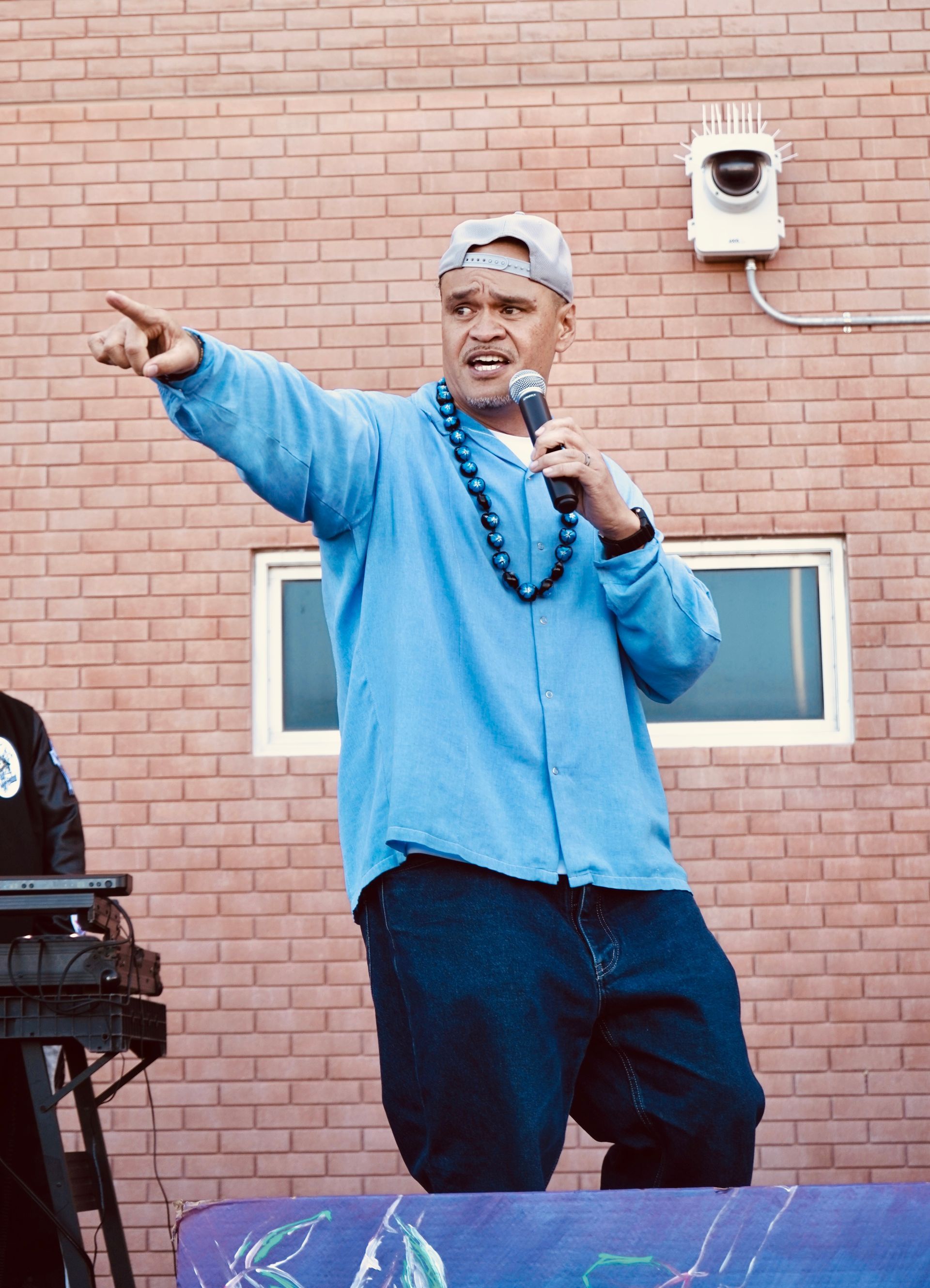 Man in a blue shirt speaking into a microphone and pointing, standing before a brick wall.