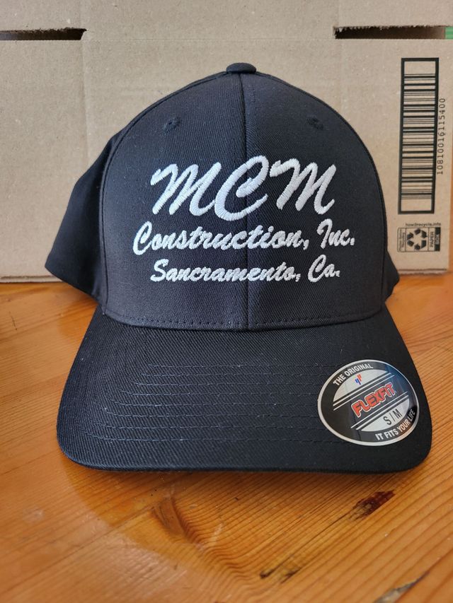 A dark blue baseball cap with white embroidered text reading "MCM Construction, Inc. Sacramento, Ca." on a wooden surface.