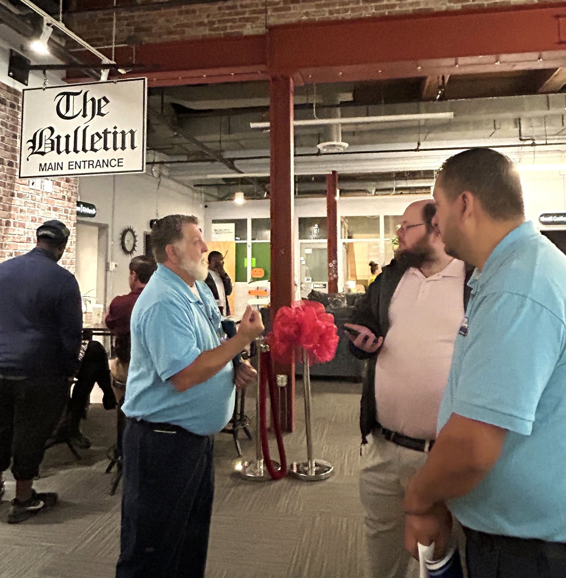 People talking in a restaurant lobby near a sign reading βThe Bulletinβ and a pink ribbon stand.