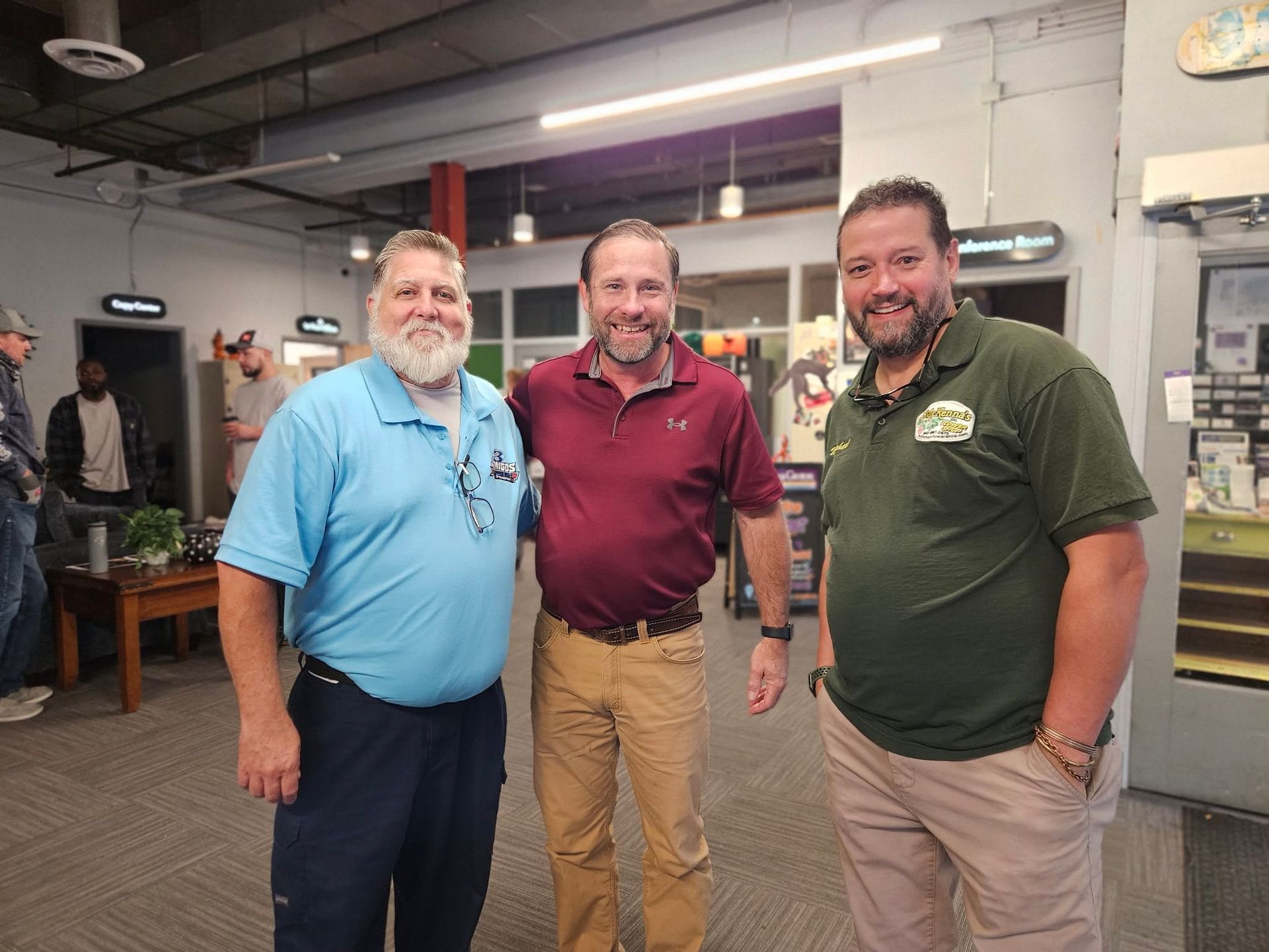 Three smiling men standing together in a workshop, wearing casual shirts and pants.