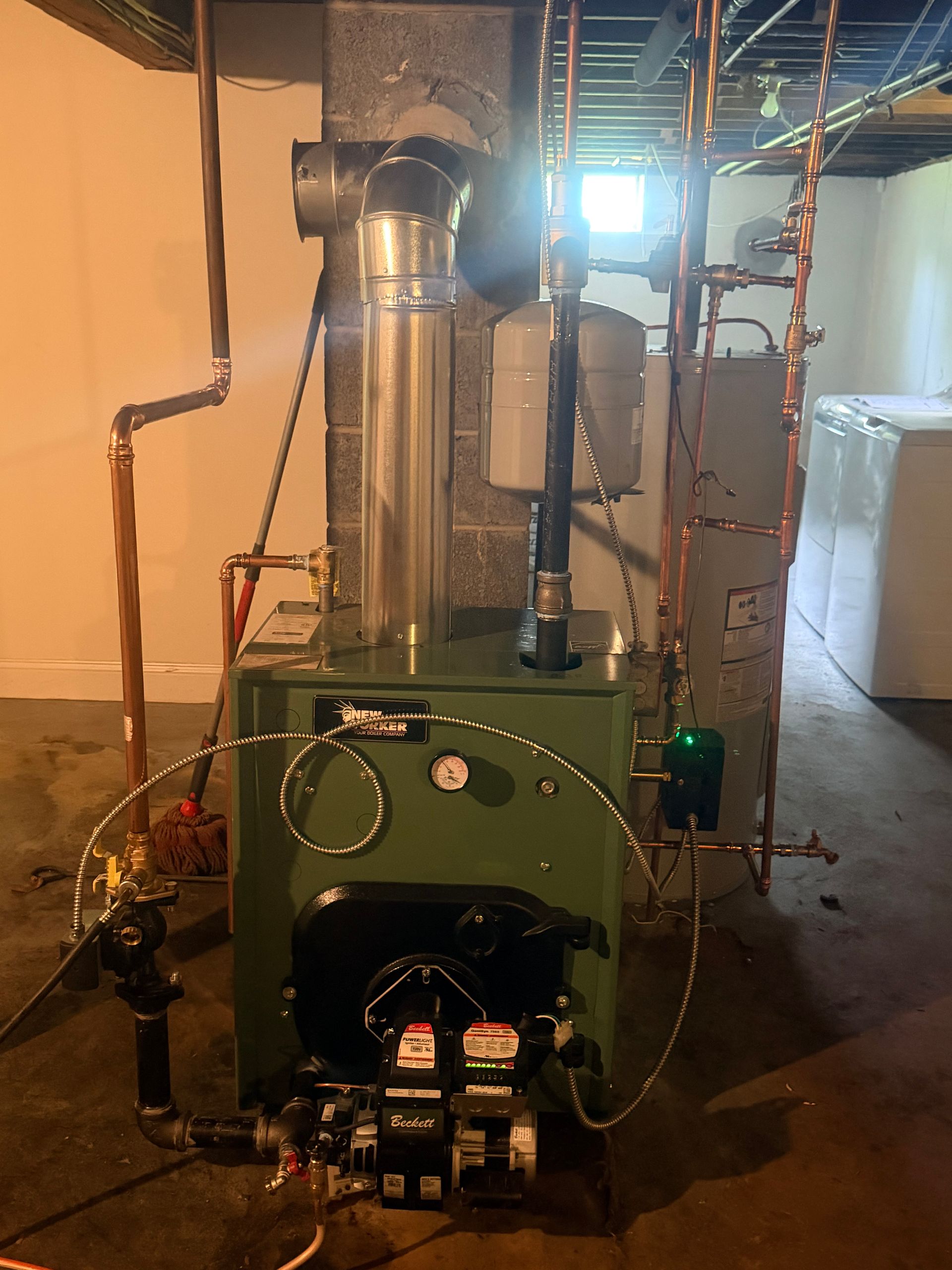 Green boiler/furnace unit in a basement, with metal vent piping, hoses, and exposed walls around it.