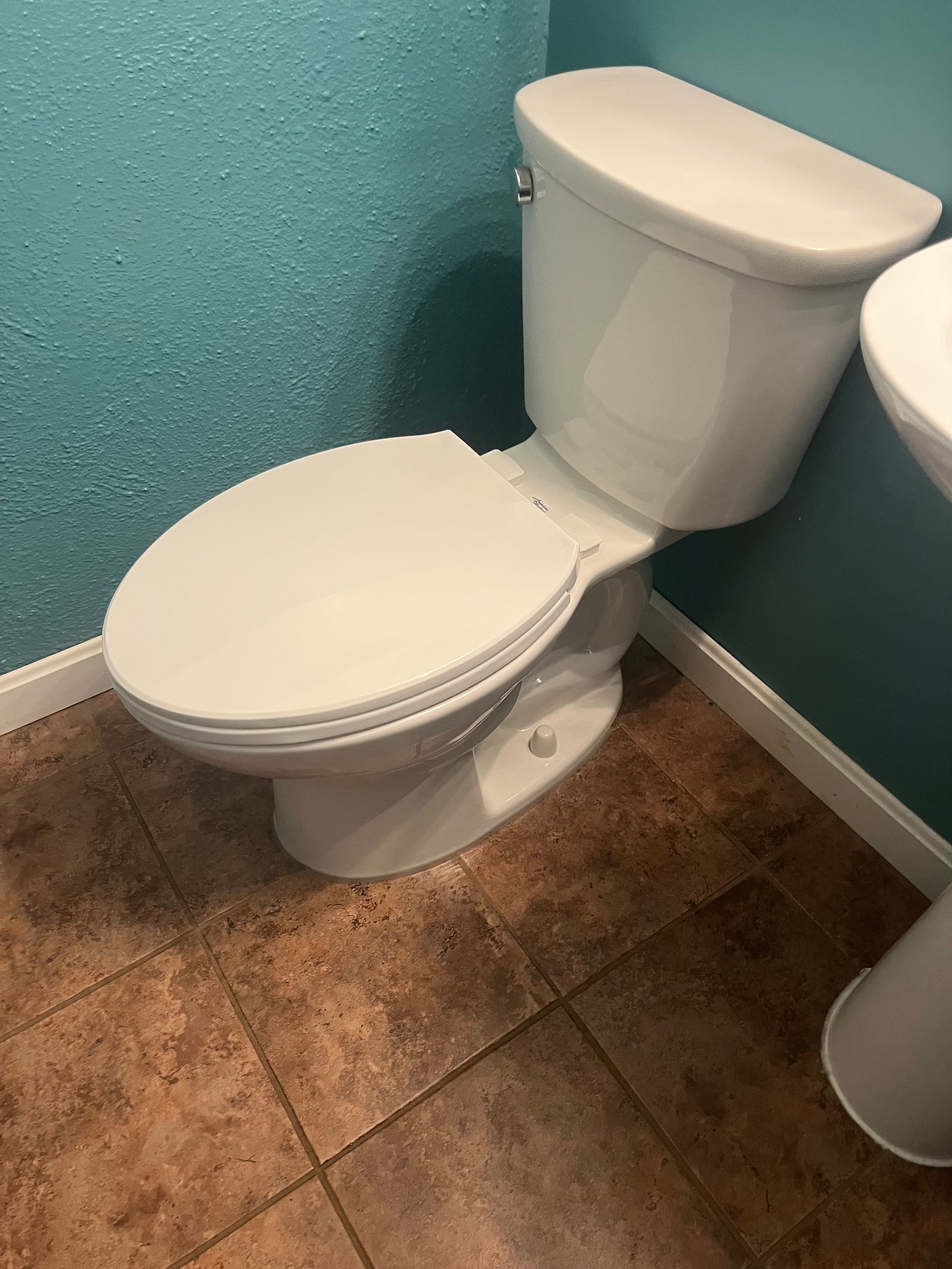 White toilet in a small bathroom with teal walls and brown tiled floor.