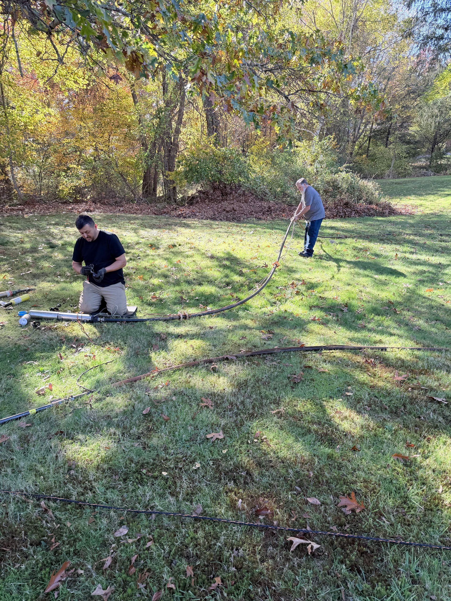 Two people working outdoors in a grassy field, one kneeling by tools and one standing nearby.