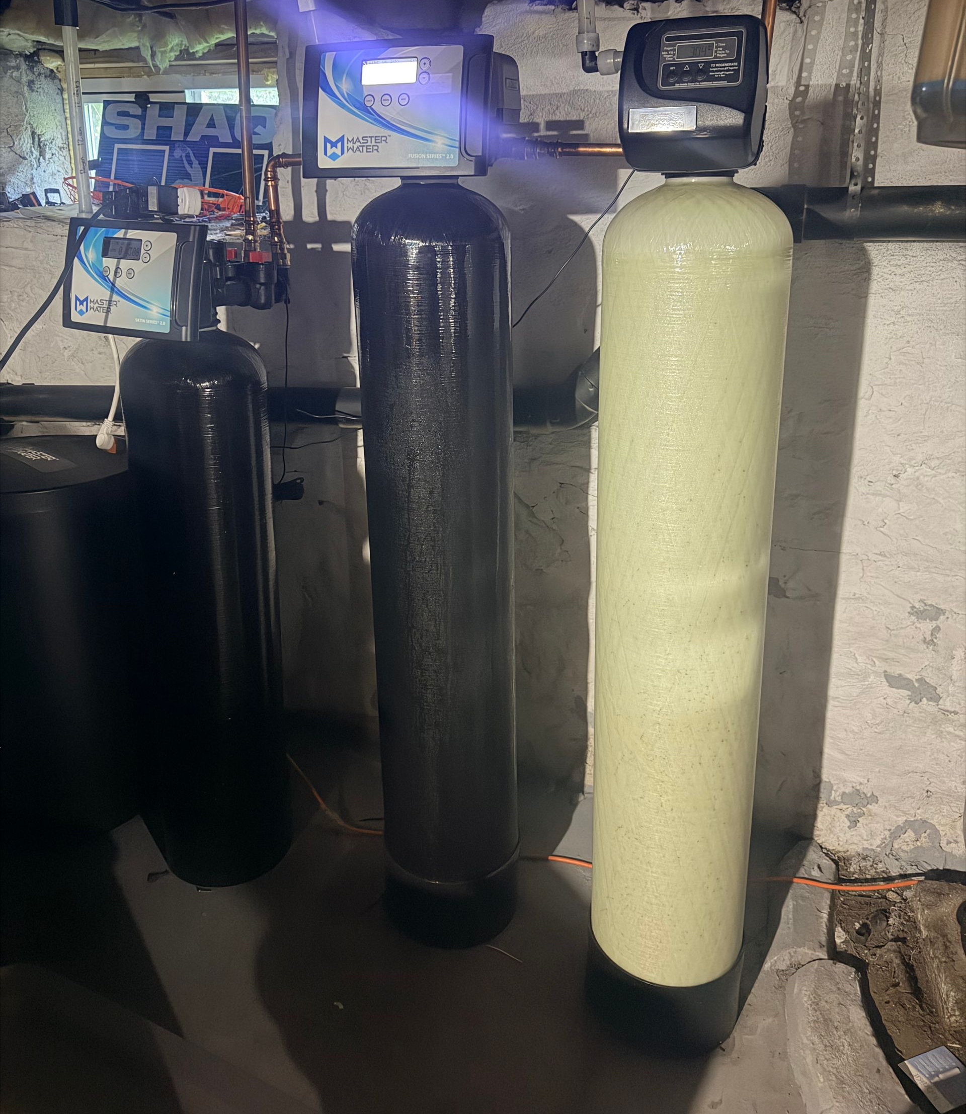 Three tall cylindrical tanks in a basement, black and cream-colored, beside pipes and utility equipment.