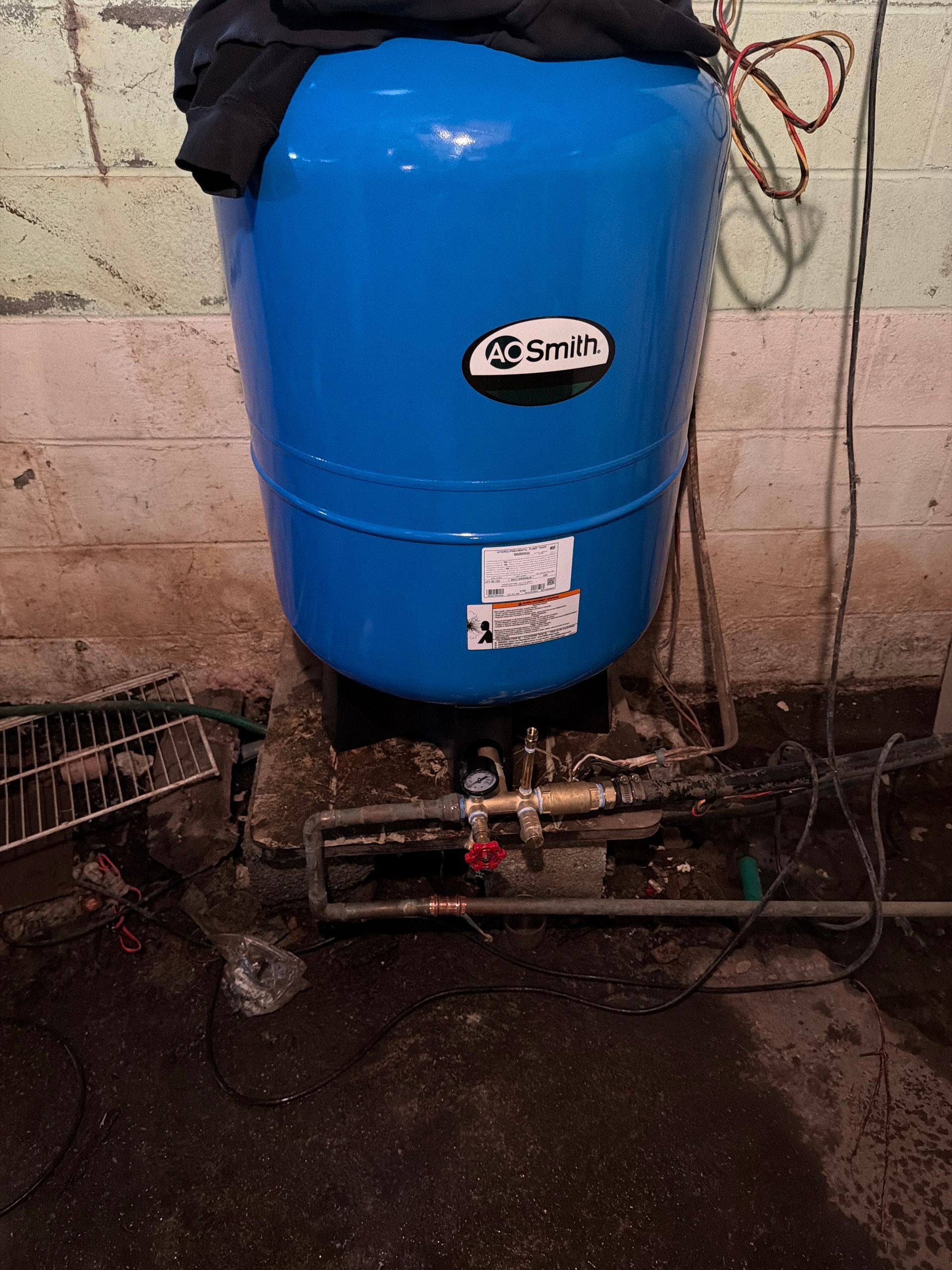 Blue water tank with pipes and valve in a tiled utility area, standing on a dark floor.