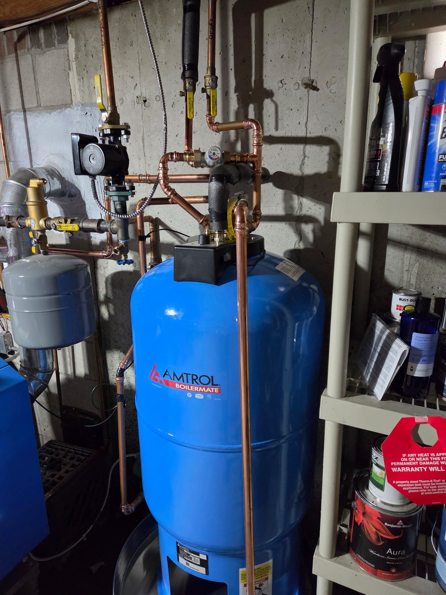 Blue boiler and copper piping in a basement utility room with a gray tank and shelves nearby
