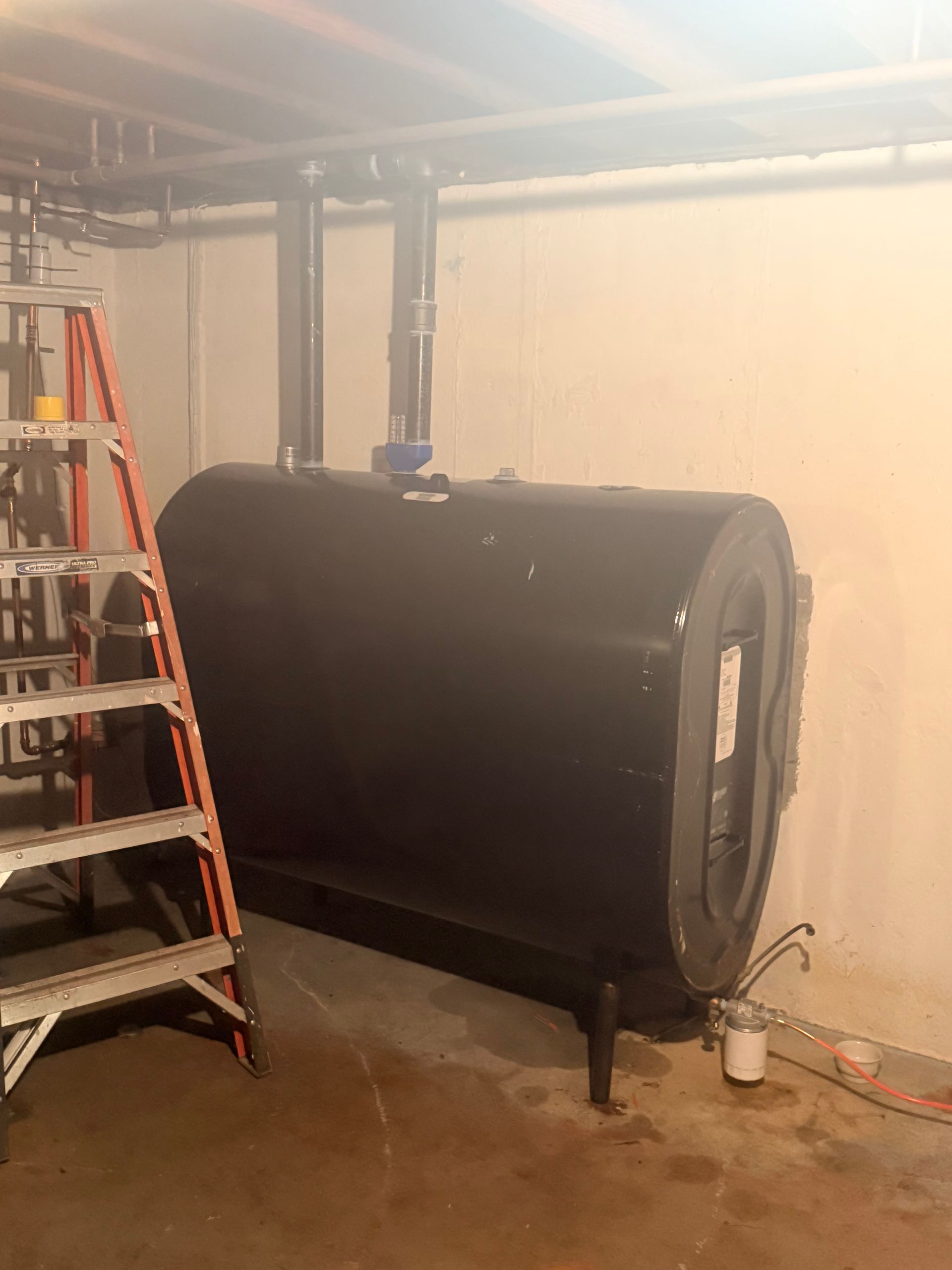 Large black cylindrical tank with pipes in a basement, beside a ladder and white wall