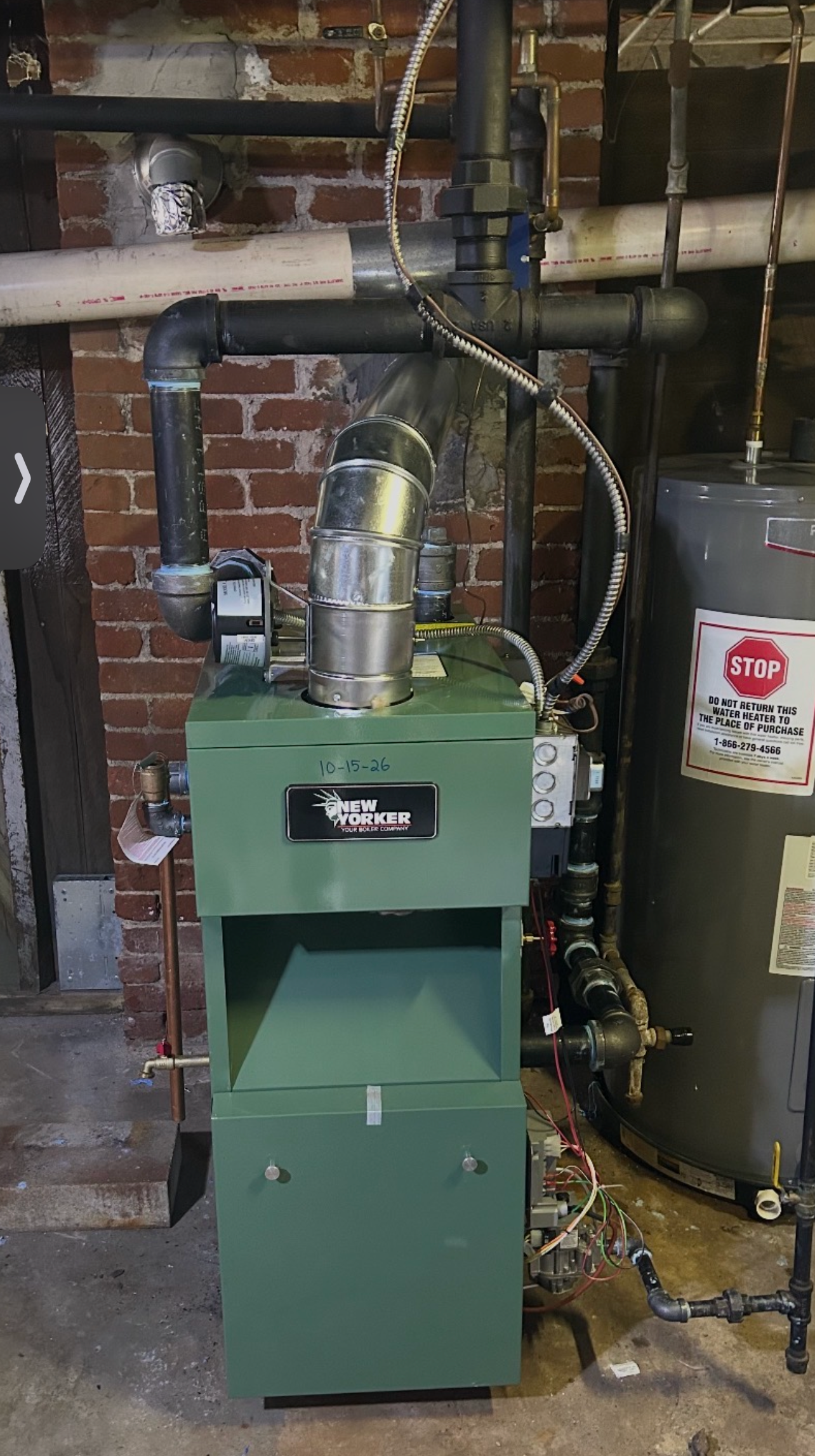 Green furnace in a basement utility room with exposed pipes and a nearby water heater